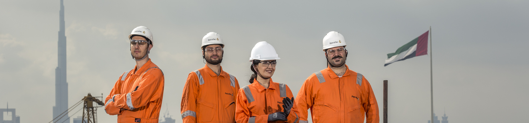 Three men and a women in orange overalls and white hard hats with Petrofac. Posing over the Dubai skyline with the Burj Khalifa in the background 